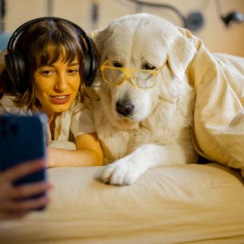 Young woman takes selfie photo or has a video call while lying with her cute dog under a blanket in bed. Spending leisure time and having fun together. Friendship with pets and coziness concept